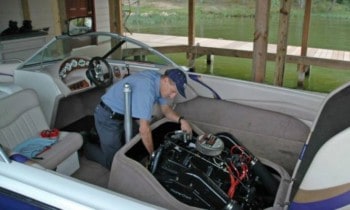 A amrine mechanic working on an inboard motor while in a baot with water and dockage in the background.