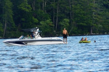 A man standing on the swim deck of his ski boat getting his kids ready to tow behind in a towable tube in the water with trees in the background.