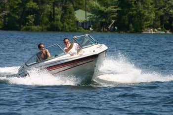 Two people in a bowrider boat driving in the water on a sunny day with trees in the background.