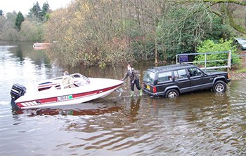 A jeep with a boat on a trailer, backed into the water at a boat ramp, ready to launch the boat with one man on the trailer toung and the other man in the boat.