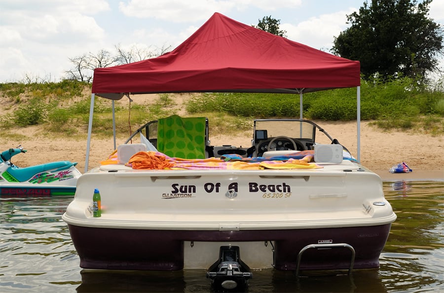 A maroon and white boat pulled up to a beach floating in the water and a pop-up canoy over the top of it kinda looking like a funny bimini top.