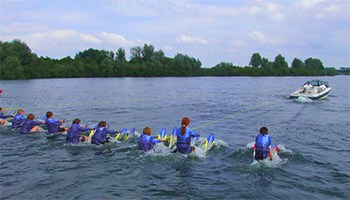 A Ski Nautique boat pulling 12 waterskiers out of the water all at once in a lake with trees in the background.