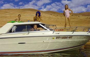 A cudy style boat with four kis climbing around it in the water with hills in the background.