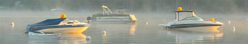 Four boats tied to mouring balls out in a lake with steam coming off the water in early morning sun.