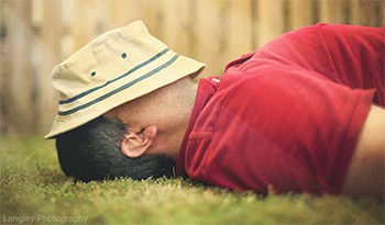 A man laying in a lawn with his hat over his face and a fence in the background on a sunny day.