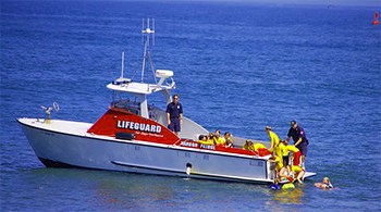 A red and white lifeguard boat out in the water with two officers and a group of children out doing some training.