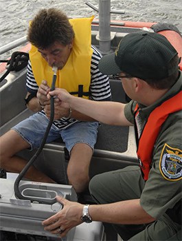 A man in a boat out on the water with his hands cuffed and an officer giving him a breathalyzer test.