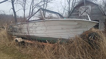 An abandoned boat on a trailer with weeds grown up all around it in front of a barn.